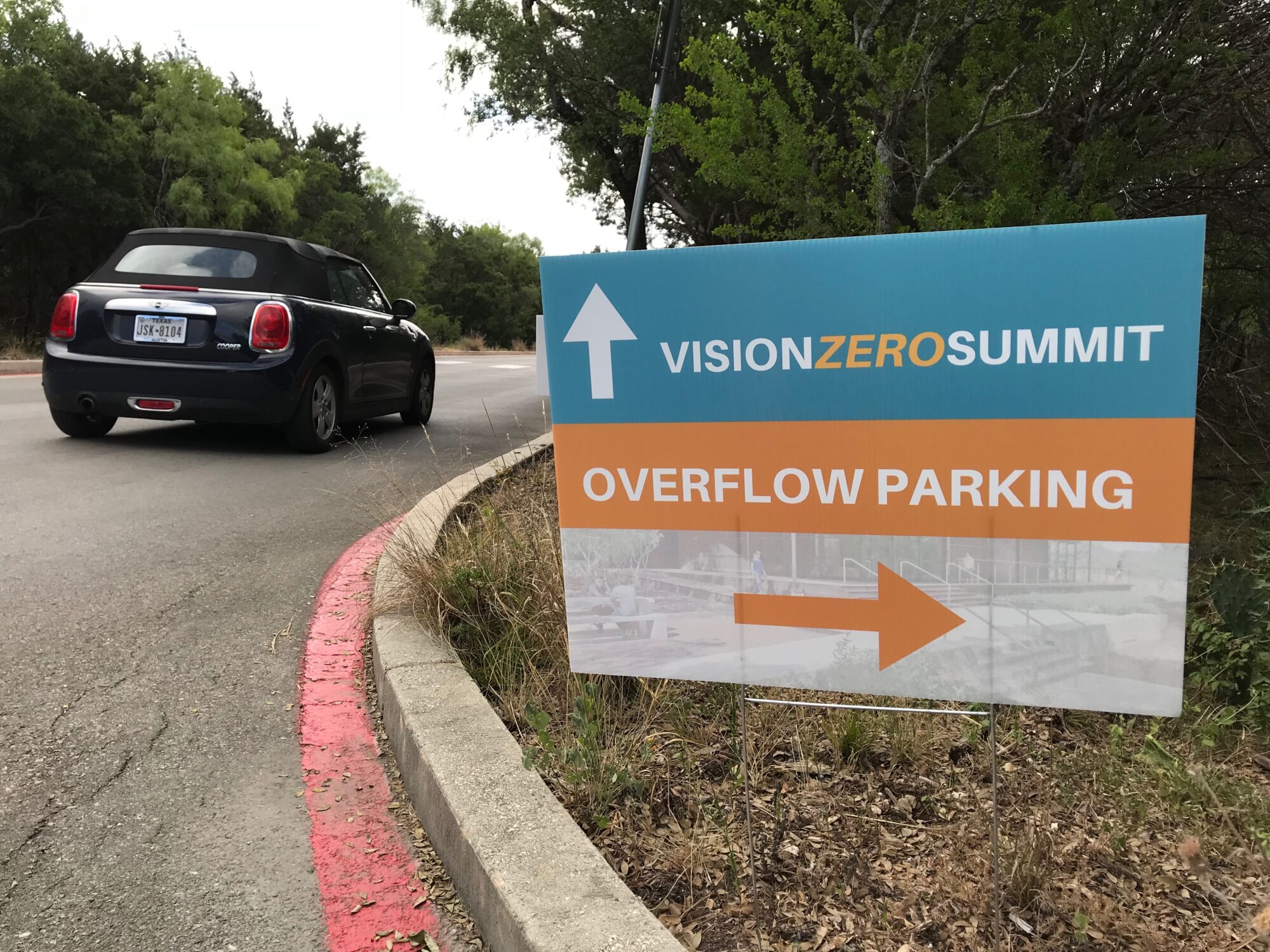 Picture of a sign on the side of a driveway that has an arrow pointing forward that says "Vision Zero Summit" and another arrow pointing to the right that says "Overflow parking"