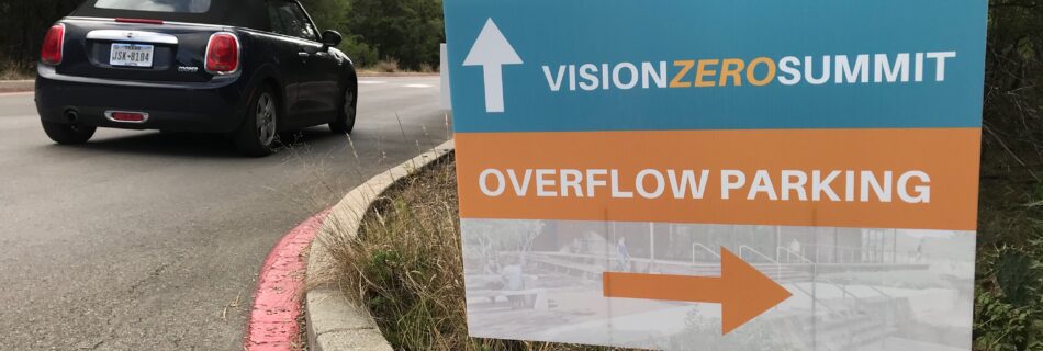 Picture of a sign on the side of a driveway that has an arrow pointing forward that says "Vision Zero Summit" and another arrow pointing to the right that says "Overflow parking"