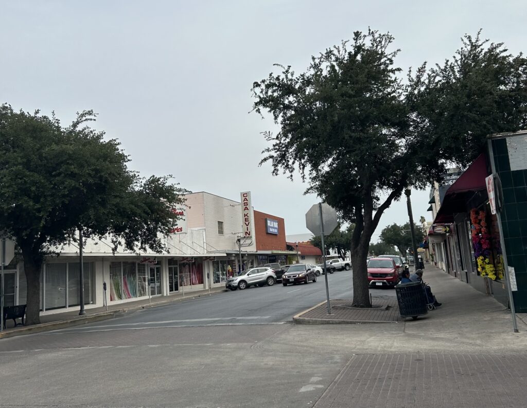 A photo of an intersection in downtown McAllen depicting raised, red brick crosswalks, curb extensions with trees, and sidewalks with benches along a shopping corridor.