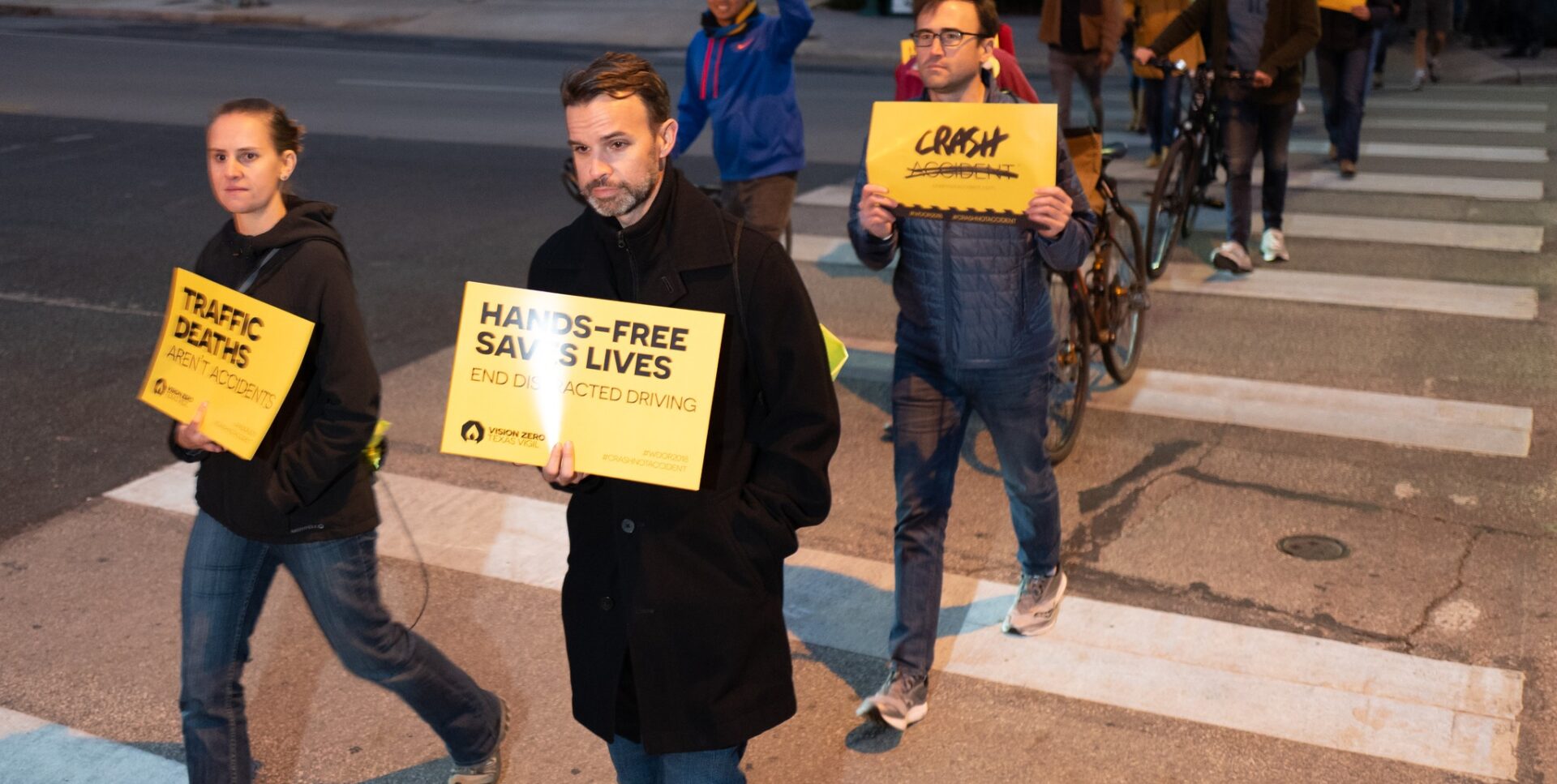 People walk across a street, carrying yellow signs that say "Hands-free saves lives" and "Crash not Accident"