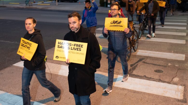 People walk across a street, carrying yellow signs that say "Hands-free saves lives" and "Crash not Accident"