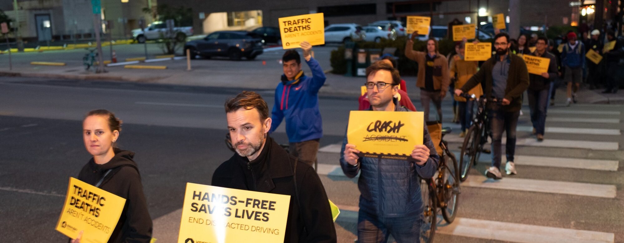 People walk across a street, carrying yellow signs that say "Hands-free saves lives" and "Crash not Accident"