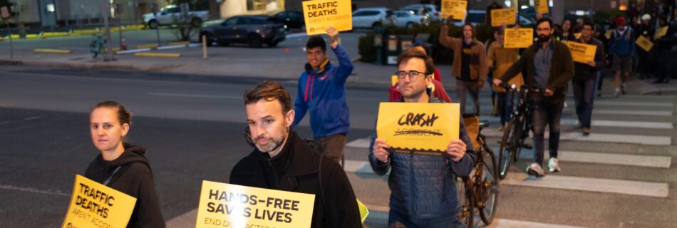 People walk across a street, carrying yellow signs that say "Hands-free saves lives" and "Crash not Accident"