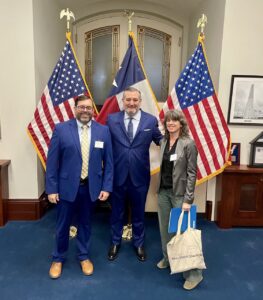 Katy Atkiss, Jay Blazek Crossley and Senator Ted Cruz pose for a photo in front of two US flags and one Texas flag.